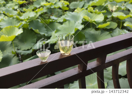 Glass teacups with Chinese tea on a wooden railing against a background of lush green lotus leaves in a garden pond 143712342