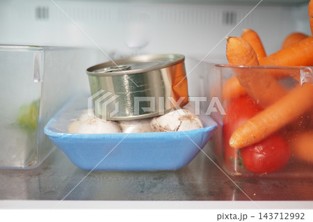 Open can resting on mushrooms beside carrots and tomatoes in a refrigerator 143712992