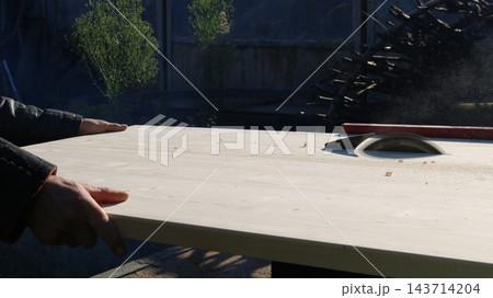 Off-camera, a carpenter carefully guides a wide beech wood panel toward the rotating saw blade of a machine. The process of rip-cutting timber takes place outdoors on a sunny day. Off-camera, a carpenter carefully guides a wide beech wood panel toward the rotating saw blade of a machine. The process of rip-cutting timber takes place outdoors on a sunny day. 143714204