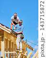 Roofing work on timber frame building involves two roofers fastening corrugated bitumen sheets with nails and hammer beside roof edge. 143715872