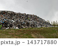Household waste is piled across a dump beside a grass field, with trees in the background under an overcast sky 143717880