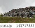 A mound of mixed waste fills a landfill beside a grassy field, with trees in the distance under an overcast sky during the day 143717882