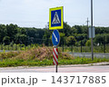 A pedestrian crossing sign and a blue direction sign stand beside a road near a fence and trees during the day 143717885