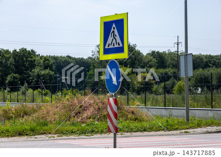 A pedestrian crossing sign and a blue direction sign stand beside a road near a fence and trees during the day 143717885