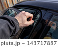 Beside a parked car on a city street, a young man opens the door during daylight and reaches toward the window frame 143717887
