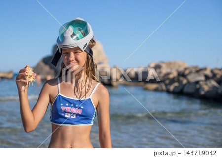Young girl wearing snorkeling mask holds seashell while standing on beach with rocky coastline and clear blue sky in the background Young girl wearing snorkeling mask holds seashell while standing on beach with rocky coastline and clear blue sky in the background 143719032