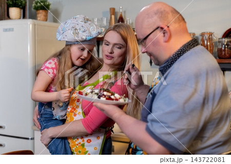 Celebratory family dessert serving moment, Family shares special dessert in cheerful kitchen, Father presents decorated treat as family gathers happily around kitchen Celebratory family dessert serving moment, Family shares special dessert in cheerful kitchen, Father presents decorated treat as family gathers happily around kitchen 143722081