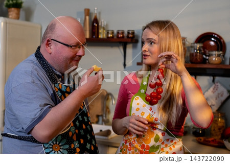Man tasting cheese sample while partner presents tomatoes, flavorfocused moment with chopping board and herbs, friendly critique and pairing discussion, warm kitchen setting 143722090
