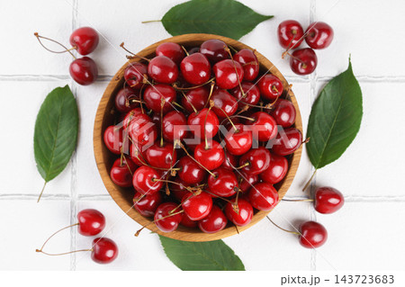 Ripe sweet cherry in wooden bowl with green leaves on white tile background, healthy organic fruit 143723683