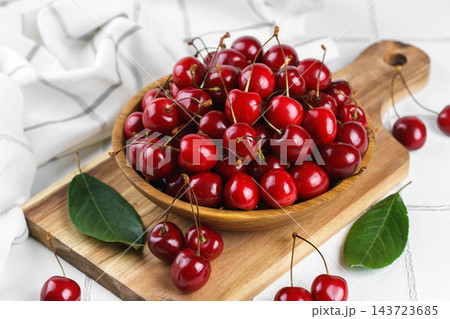 Fresh ripe sweet cherry in wooden bowl on rustic cutting board with green leaves on white background Fresh ripe sweet cherry in wooden bowl on rustic cutting board with green leaves on white background 143723685