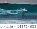 Surfer man crouching low on a white surfboard while carving through a deep blue wave with ocean spray 143724633