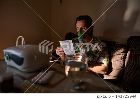 Man using oxygen mask and home nebulizer reading instructions with medication on a dim living room table Man using oxygen mask and home nebulizer reading instructions with medication on a dim living room table 143725944
