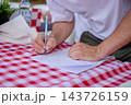A person writes on a sheet of paper at a red and white checkered table outdoors during the day 143726159