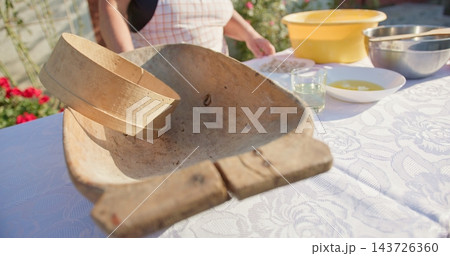 Old wooden dough trough with round wooden sieve on white patterned tablecloth outdoors under warm daylight lighting filling foreground with baking bowls and utensils in background Old wooden dough trough with round wooden sieve on white patterned tablecloth outdoors under warm daylight lighting filling foreground with baking bowls and utensils in background 143726360