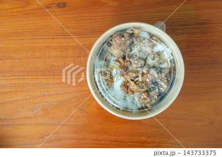 Grilled meat and rice topped with sesame seeds in a round takeout container on a wooden table. High angle view of a convenient packaged meal. 143733375
