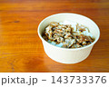 Grilled meat over white rice topped with sesame seeds, served in a disposable paper bowl on a wooden tabletop. This takeout-style meal represents fast-casual Asian cuisine. 143733376