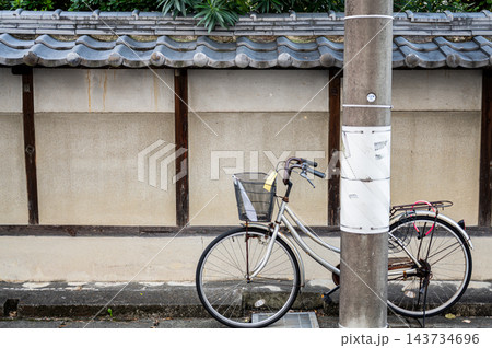 瓦屋根と白い塀が続く住宅街の路地脇に停められた銀色の自転車と電柱、前かごや荷台が見える日本の日常風景 瓦屋根と白い塀が続く住宅街の路地脇に停められた銀色の自転車と電柱、前かごや荷台が見える日本の日常風景 143734696