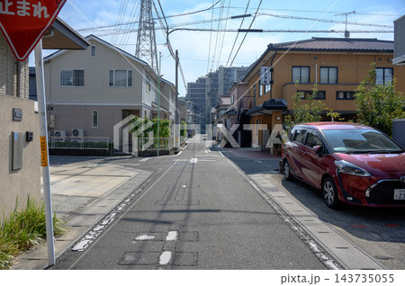 赤い車が停まる住宅街の細い生活道路と一時停止標識、電柱や電線、鉄塔と住宅が並ぶ晴れた日本の街角の風景 赤い車が停まる住宅街の細い生活道路と一時停止標識、電柱や電線、鉄塔と住宅が並ぶ晴れた日本の街角の風景 143735055