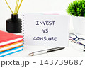 A notebook reads invest versus consume beside stacked notebooks, a pen, glasses, and a plant on a desk in a bright indoor setting 143739687
