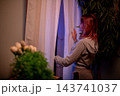 Woman with red hair reads outdoors. Caucasian woman with red hair looking through window at dusk. Quiet evening scene with woman in cozy apartment reading on her phone Woman with red hair reads outdoors. Caucasian woman with red hair looking through window at dusk. Quiet evening scene with woman in cozy apartment reading on her phone 143741037