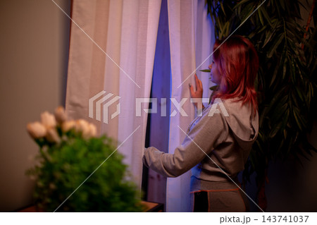 Woman with red hair reads outdoors. Caucasian woman with red hair looking through window at dusk. Quiet evening scene with woman in cozy apartment reading on her phone 143741037