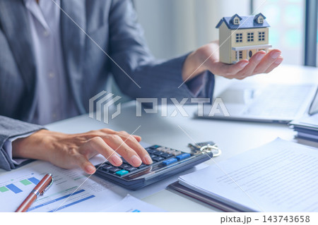 Real estate agent calculating mortgage loan costs with house model and contract on desk. Real estate agent calculating mortgage loan costs with house model and contract on desk. 143743658