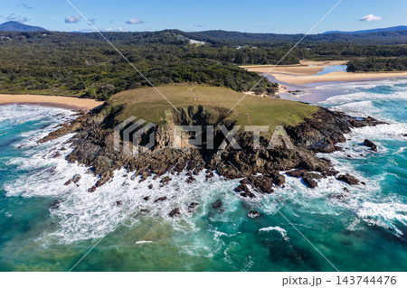 Aerial view of waves breaking around a rocky headland at Sapphire Beach, with a grassy bluff, turquoise water and sandy shoreline near Coffs Harbour, Australia. 143744476