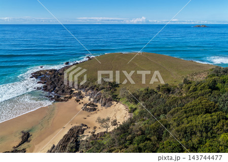 Aerial view across a grassy headland at Sapphire Beach, with rocky shoreline, breaking surf, a sandy cove and a distant offshore island near Coffs Harbour. Aerial view across a grassy headland at Sapphire Beach, with rocky shoreline, breaking surf, a sandy cove and a distant offshore island near Coffs Harbour. 143744477