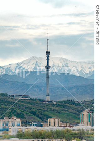 Tall red and white television tower rising above Almaty city buildings with snowy mountains behind 143748425