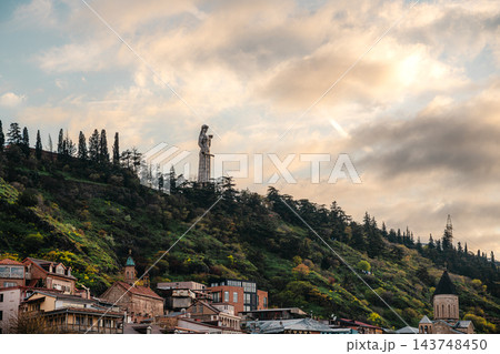 Mother of Georgia statue on a green hillside above old town houses under golden sunset clouds in Tbilisi 143748450