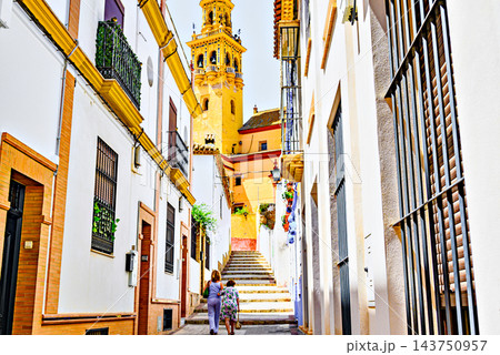 Two women walk up the street leading to the Church of Santiago in Alcala de Guadaira, with the church's bell tower in the background; Seville. Two women walk up the street leading to the Church of Santiago in Alcala de Guadaira, with the church's bell tower in the background; Seville. 143750957