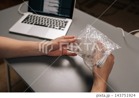 Hands peeling protective film from white laptop charger, representing beginning of setup or unboxing new electronic device for immediate use at a workstation. Hands peeling protective film from white laptop charger, representing beginning of setup or unboxing new electronic device for immediate use at a workstation. 143751924