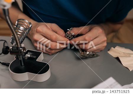 Detail cropped shot of technician male working on electronics using a soldering iron and a magnifying lamp, carefully assembling or repairing a circuit board at a workbench. Detail cropped shot of technician male working on electronics using a soldering iron and a magnifying lamp, carefully assembling or repairing a circuit board at a workbench. 143751925