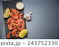 There are frozen shrimps and two lemon slices on a black stone board, and a salt shaker and a head of garlic are placed next to it on a gray background. Concept: a minimalistic set of ingredients 143752330