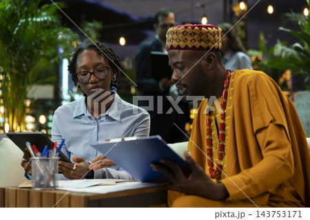 Business partners compare information on tablet and printed documents during discussion at night. Professionals crosschecking data between device and paperwork on clipboard in rooftop workspace 143753171