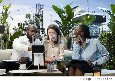 Businessman points to tablet screen during rooftop lounge workspace presentation with coworkers. Company team analyzing project information on device during meeting on rooftop terrace 143753172