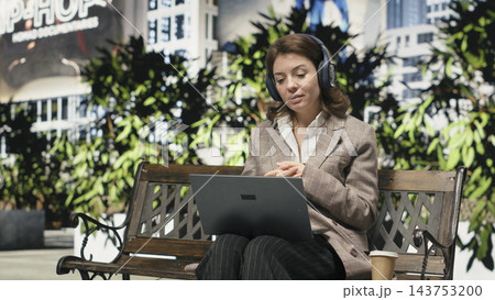Businesswoman works remotely from park bench, having video conference on laptop. Woman wearing headphones communicates with colleagues online, enjoying flexible outdoor workplace Businesswoman works remotely from park bench, having video conference on laptop. Woman wearing headphones communicates with colleagues online, enjoying flexible outdoor workplace 143753200