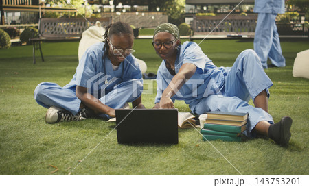 Women students team in blue scrubs work together with laptop and textbooks, group study on campus grass with stacked books. Outdoor learning and med school study session. 143753201