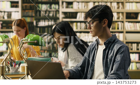 Laptop, student and studying as young man with glasses works on a computer beside fellow readers. Research, digital learning and academic productivity support coursework in a library. Laptop, student and studying as young man with glasses works on a computer beside fellow readers. Research, digital learning and academic productivity support coursework in a library. 143753273