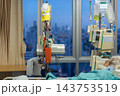 Infusion bags hang beside electronic medical pumps near hospital bed green bedding patient partly visible while large window reveals distant city view cool evening light 143753519