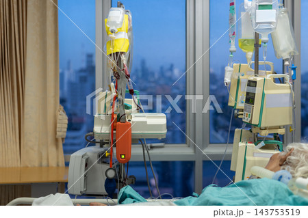 Infusion bags hang beside electronic medical pumps near hospital bed green bedding patient partly visible while large window reveals distant city view cool evening light Infusion bags hang beside electronic medical pumps near hospital bed green bedding patient partly visible while large window reveals distant city view cool evening light 143753519