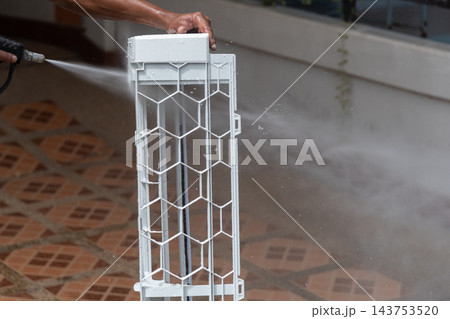 A person holds white air conditioner filter frame upright while spraying it water wet tiled floor mesh panel geometric protective grid visible the cleaning view A person holds white air conditioner filter frame upright while spraying it water wet tiled floor mesh panel geometric protective grid visible the cleaning view 143753520