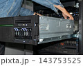 A technician denim sleeve works metal server unit extended from an equipment rack drive bays orange release tabs cables visible close view of hardware maintenance 143753525