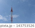 A slender red white lattice communications tower rises against pale blue sky clouds antenna equipment near the top leaving broad open space around the tall metal structure 143753526