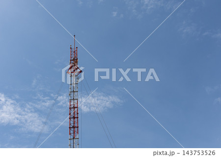 A slender red white lattice communications tower rises against pale blue sky clouds antenna equipment near the top leaving broad open space around the tall metal structure A slender red white lattice communications tower rises against pale blue sky clouds antenna equipment near the top leaving broad open space around the tall metal structure 143753526