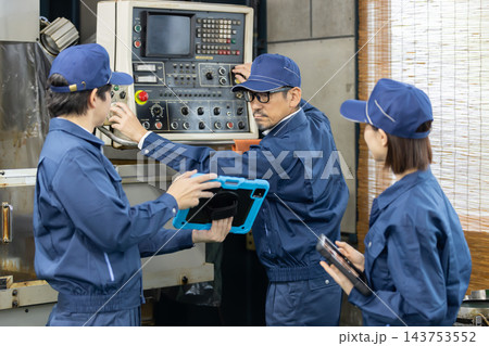 製造工場で工作機械の操作方法を若手男女従業員に指導するベテラン男性技術者 製造工場で工作機械の操作方法を若手男女従業員に指導するベテラン男性技術者 143753552