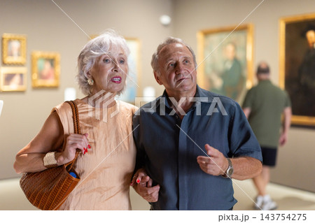 Senior couple walk around museum hall, behind they was variety of paintings Senior couple walk around museum hall, behind they was variety of paintings 143754275