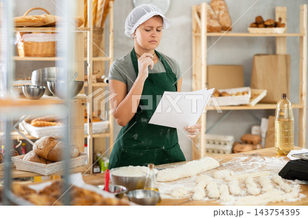 Young female baker read a new recipe on paper in the kitchen of private bakery Young female baker read a new recipe on paper in the kitchen of private bakery 143754395