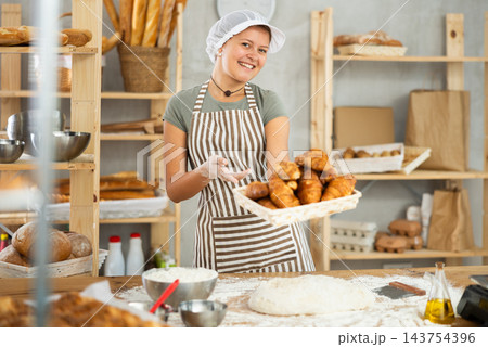 Smiling female baker in uniform stands behind counter in small private bakery offering croissants Smiling female baker in uniform stands behind counter in small private bakery offering croissants 143754396