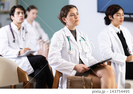 Young girl medical student dressed in labcoat sitting and listening lecture, taking notes on tablet in lecture hall Young girl medical student dressed in labcoat sitting and listening lecture, taking notes on tablet in lecture hall 143754495